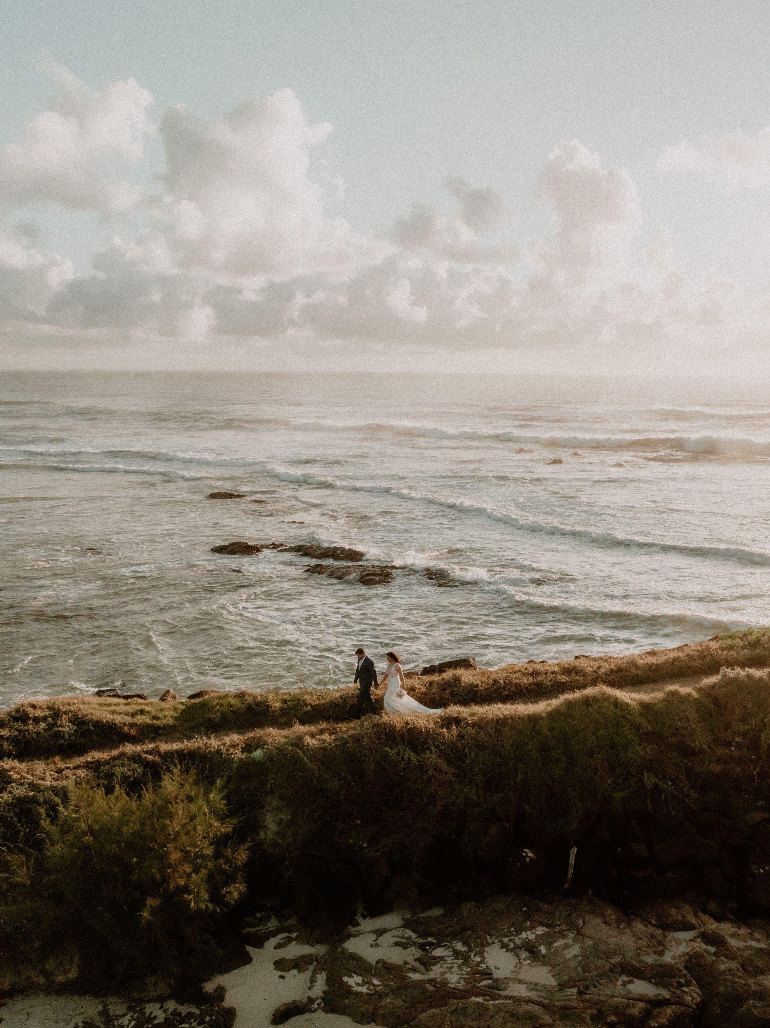 Bride and Groom walking together in a dirt road in front of the ocean during sunset