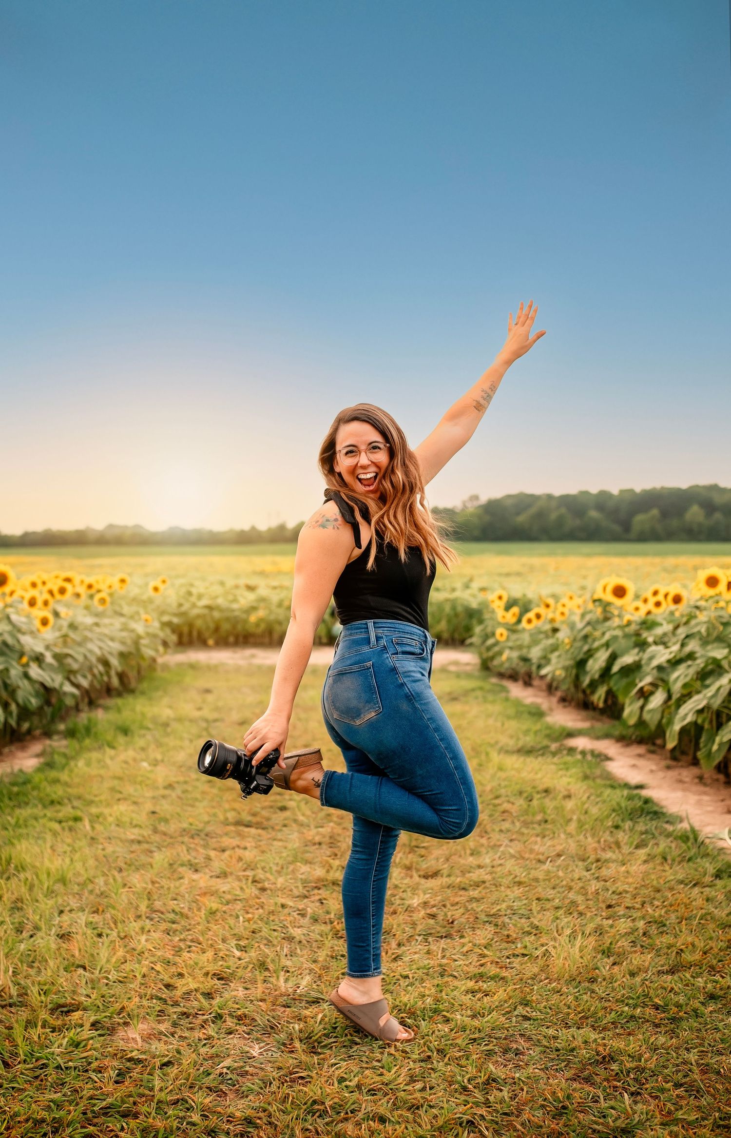 Joyful photographer in denim jumping with camera in sunflower field at sunset.
