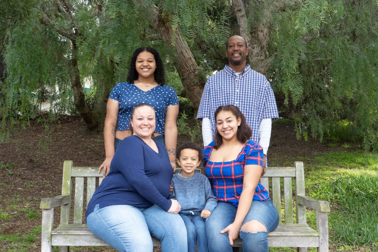 Happy family of five posing together on a wooden bench surrounded by lush greenery.