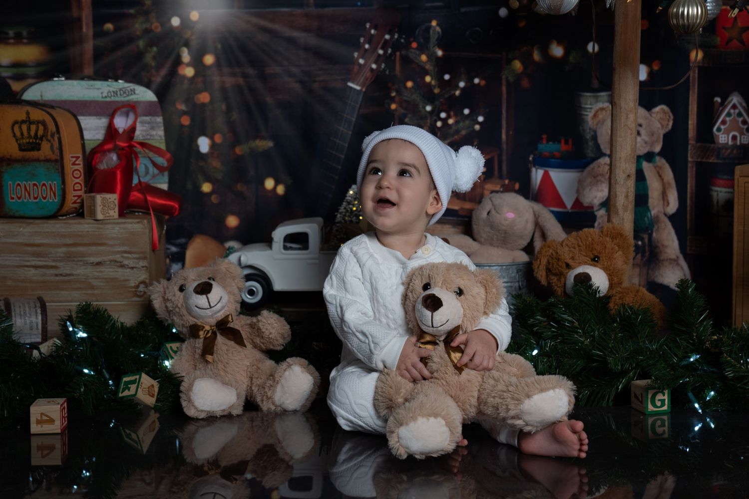 A toddler in white sits with plush teddy bears amid Christmas decorations and twinkling lights.
