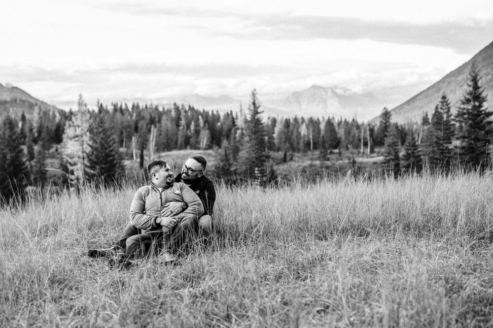 couple sitting in a grassy field with a mountain backdrop, looking at each other smiling