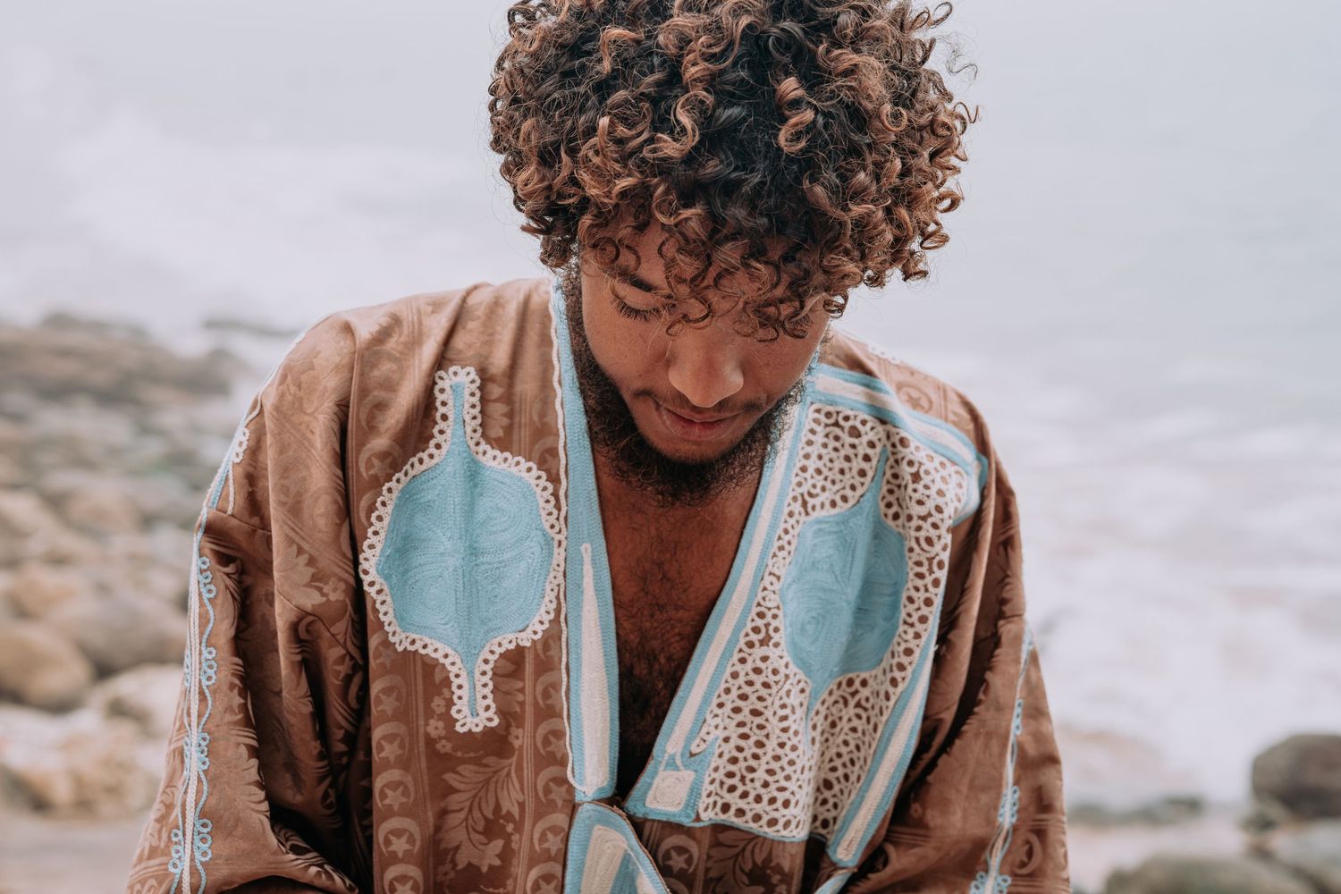 Person wearing an ornate brown and turquoise patterned robe against a cloudy coastal backdrop.