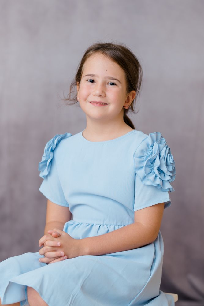 Child wearing a light blue dress with ruffled sleeves poses for portraits against a gray backdrop.