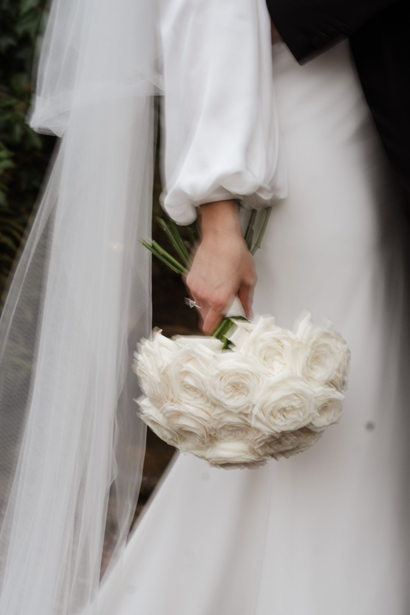 A bride in a white wedding dress holds an elegant bouquet of white roses against flowing fabric.