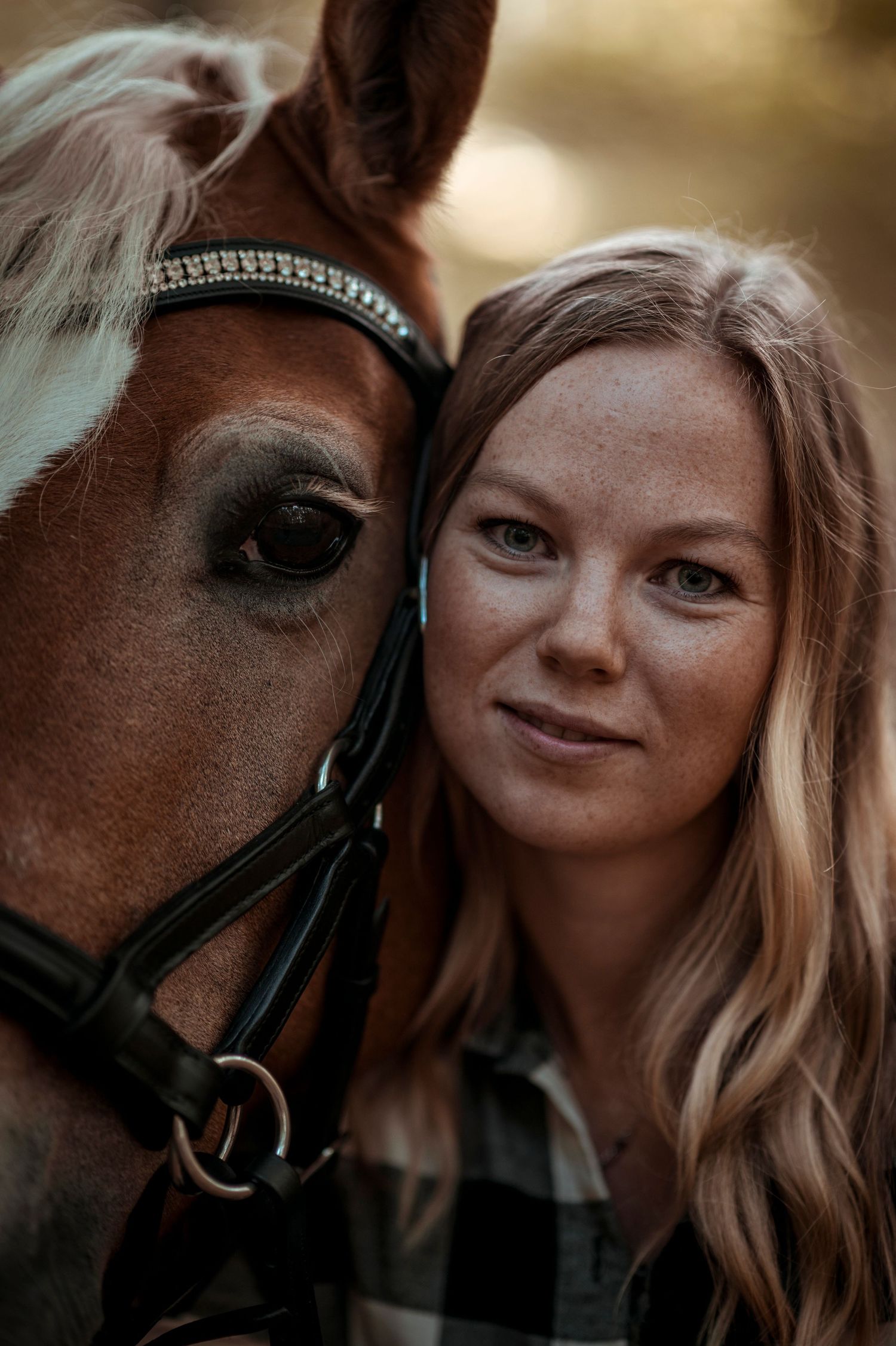 Young woman with freckles posing cheek-to-cheek with a chestnut horse wearing a bridle outdoors.