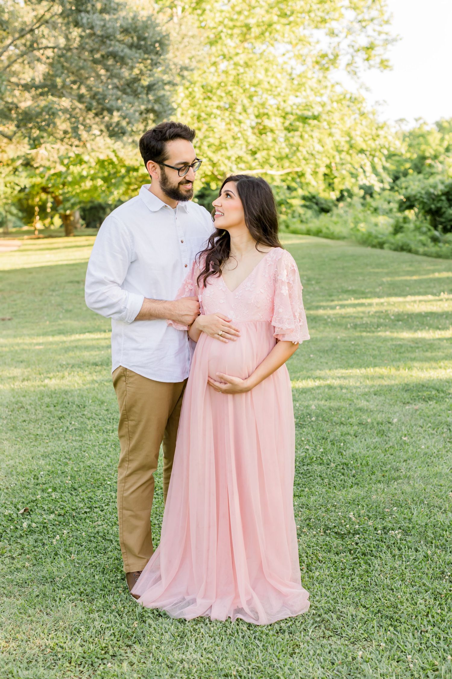 Couple embraces in sunlit outdoor setting with woman in flowing pink maternity gown.