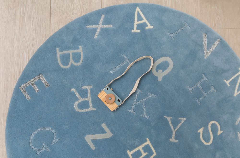 Blue round alphabet letter rug on wooden floor with small toy camera accessory.