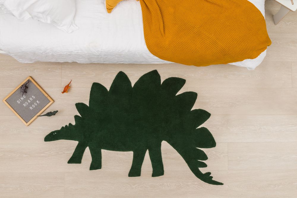 Dark green dinosaur-shaped stegosaurus rug beside bed with mustard blanket and letter board.