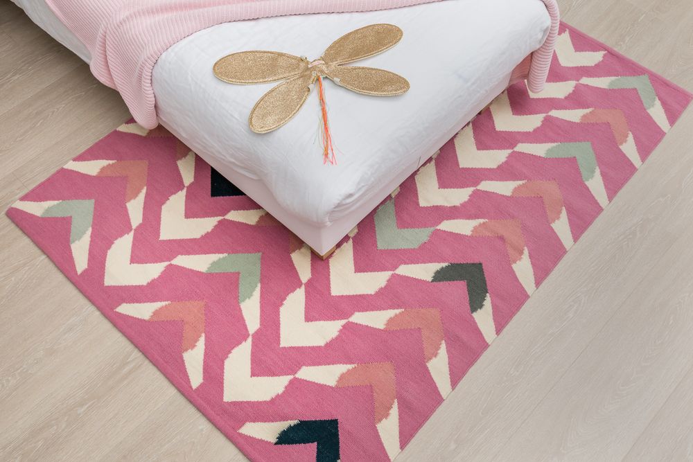 Pink chevron zigzag patterned rug with multicolor arrows under a bed with a gold dragonfly decoration.