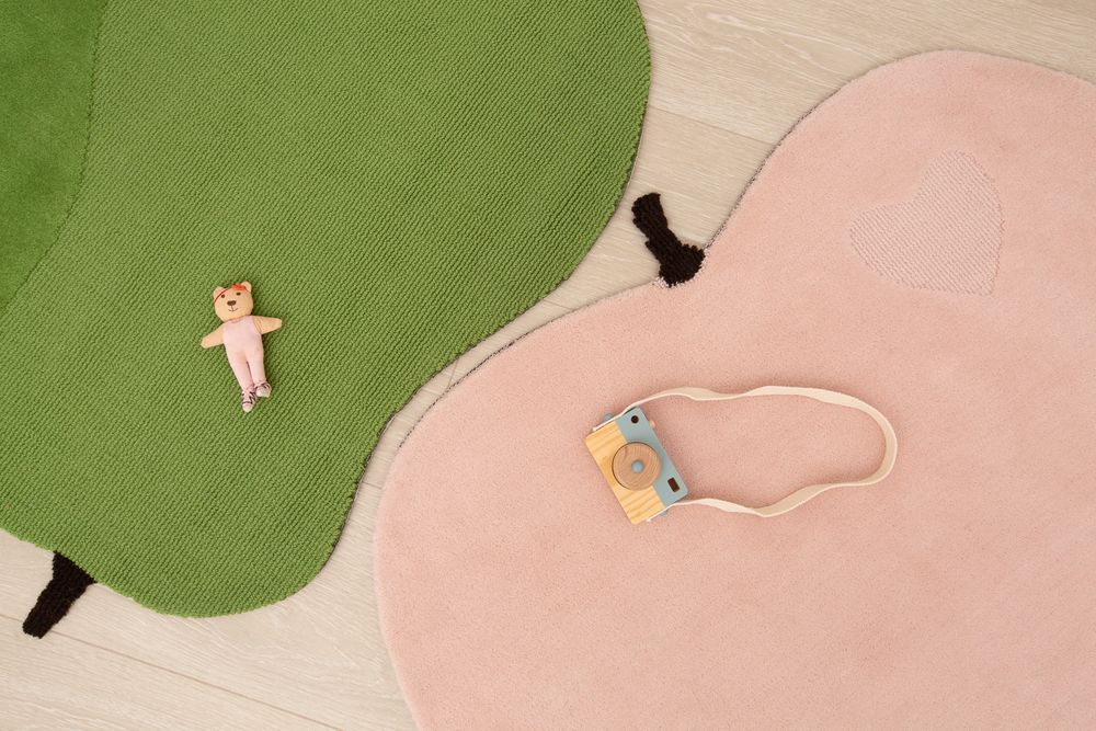 Two shaped rugs in green and pink on light wood floor with small toy bear and wooden camera toy.