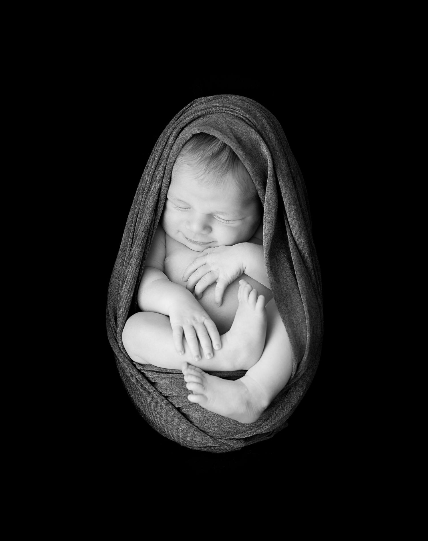 Artistic black and white newborn photography with baby wrapped in dark fabric.
