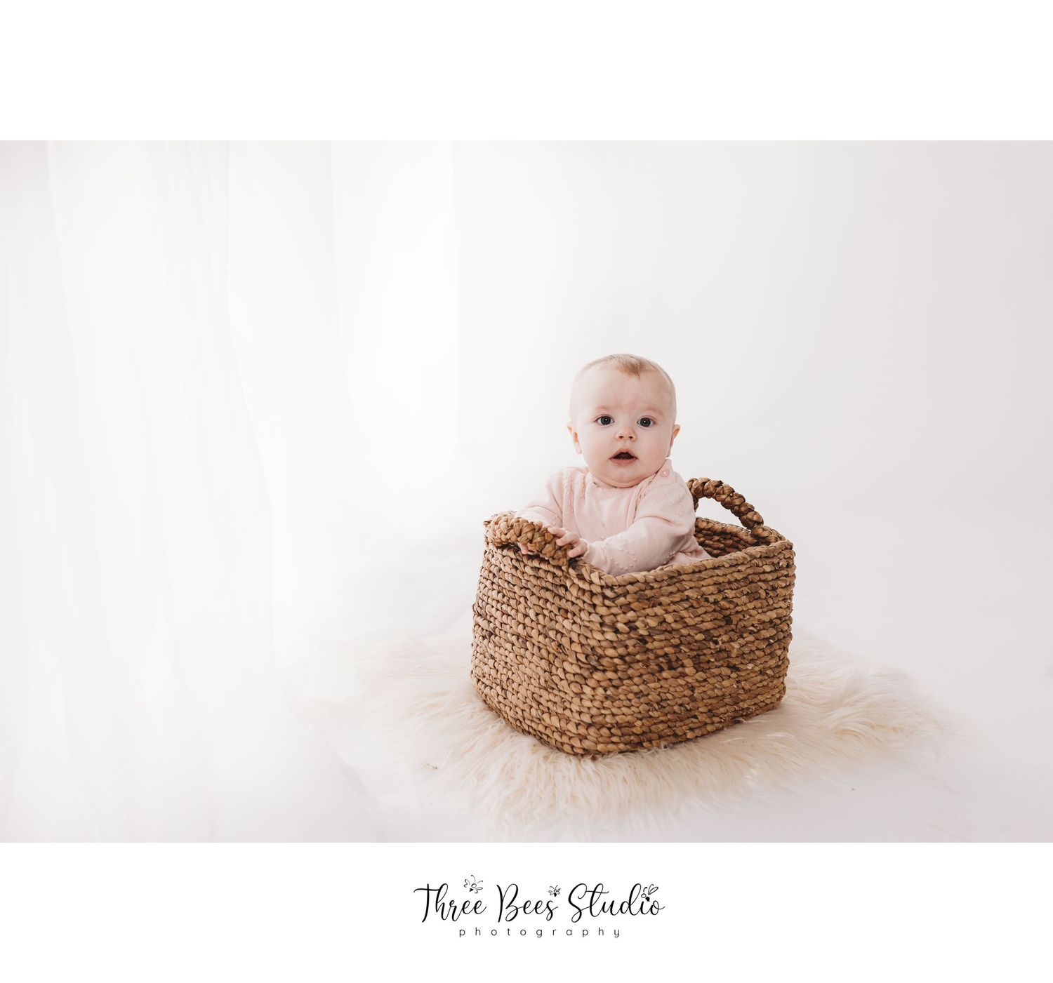 Baby sitting in woven basket against white studio backdrop.