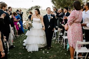 Outdoor wedding ceremony with guests seated on white chairs under trees.