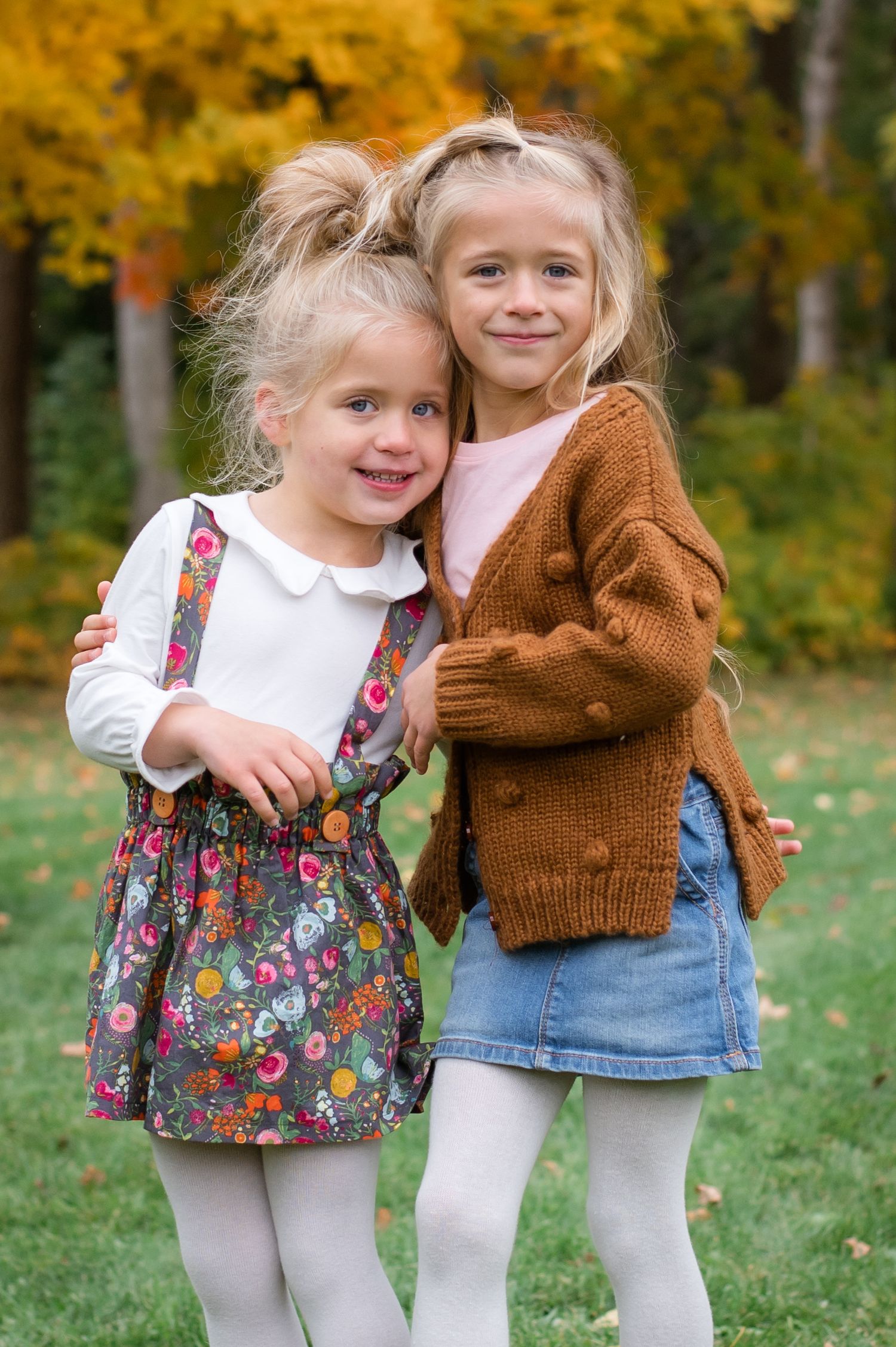 Two children in autumn attire stand together against a backdrop of golden fall leaves.