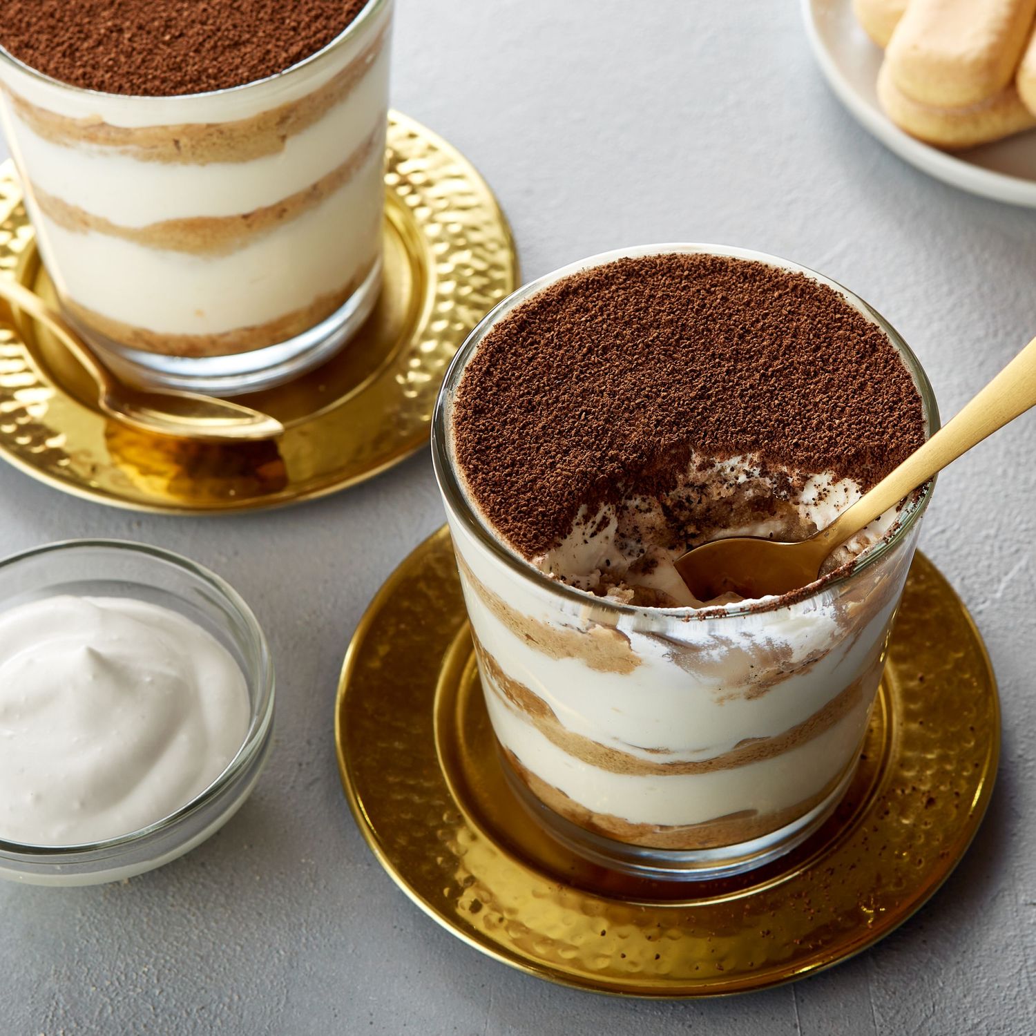 Individual tiramisu desserts topped with cocoa powder served in elegant glass cups on gold saucer plates.