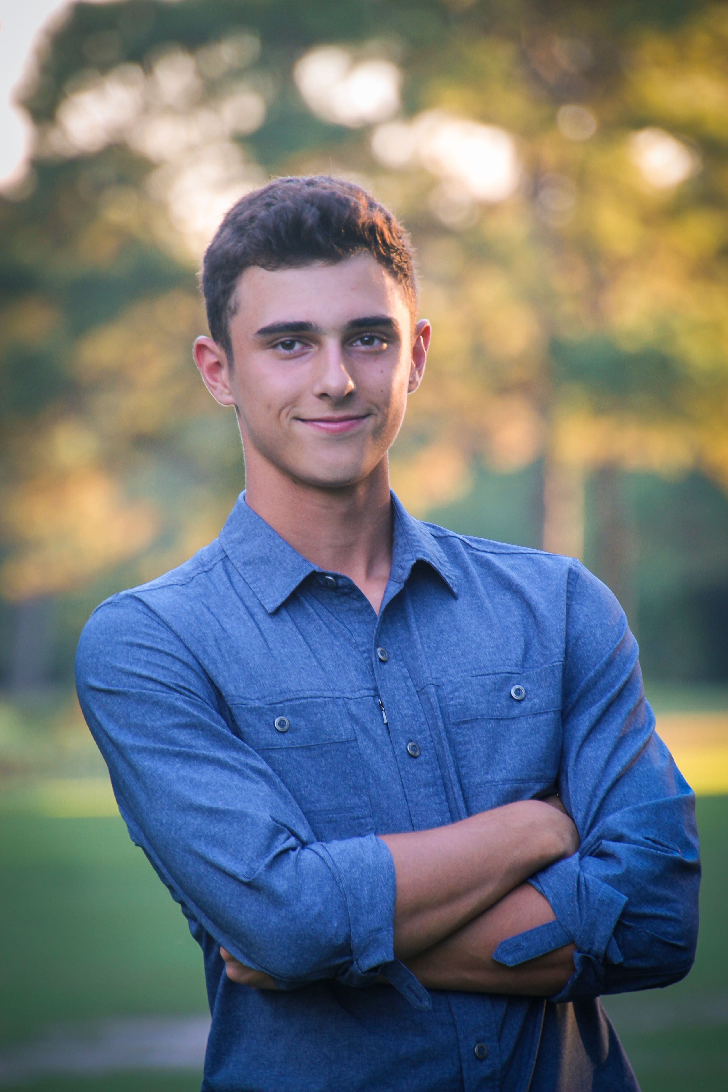 Professional outdoor portrait of a young man in blue button-down shirt with arms crossed.