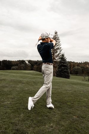 Golfer in navy shirt and khakis mid-swing on course with evergreen trees.