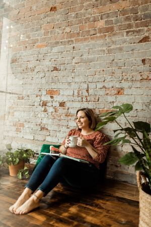 Person relaxing in a cozy corner with houseplants against exposed brick wall, reading from a book.