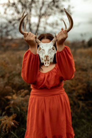 Someone holding up a decorative deer skull while wearing a rust-colored dress in an autumn field.