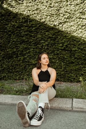 Woman in black top sitting by ivy-covered wall wearing casual attire.