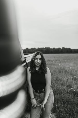 Black and white portrait leaning against car in rural field setting.