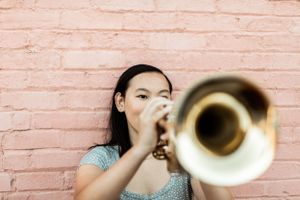 Musician playing brass trumpet against pink brick wall in casual attire.