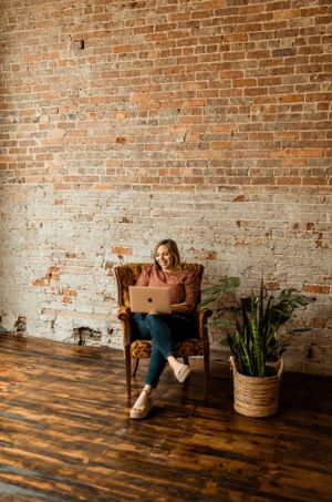 Woman works on laptop while relaxing in vintage armchair next to potted plants and brick wall.