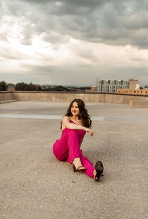 Person in bright fuchsia outfit sits playfully on concrete rooftop with cityscape in background.