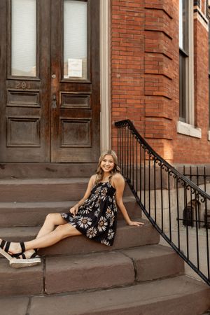 Woman in floral dress sitting on brownstone steps with wrought iron railing.