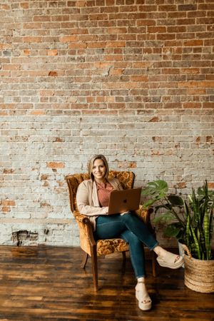 Modern workspace scene with comfortable armchair, laptop, and plants against rustic brick wall.