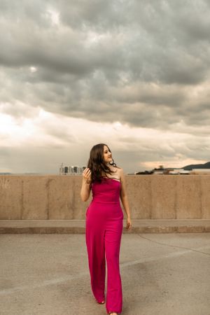 Woman wearing long pink jumpsuit smiles while walking on rooftop under dramatic cloudy sky.