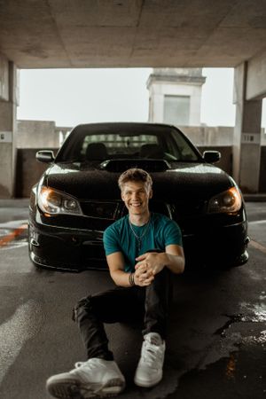 Car enthusiast poses with black sports car in covered parking garage.