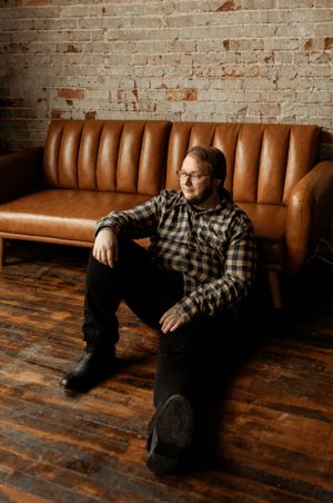 Casual indoor portrait on brown leather couch with brick wall background.