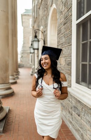 Graduate in white dress and cap smiling in front of stone pillars.