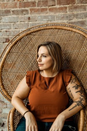 Figure seated in woven peacock chair wearing rust-colored top against brick wall.