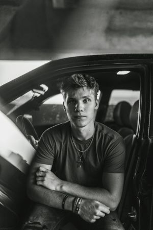 Black and white portrait of someone sitting in a car wearing a t-shirt and necklace.