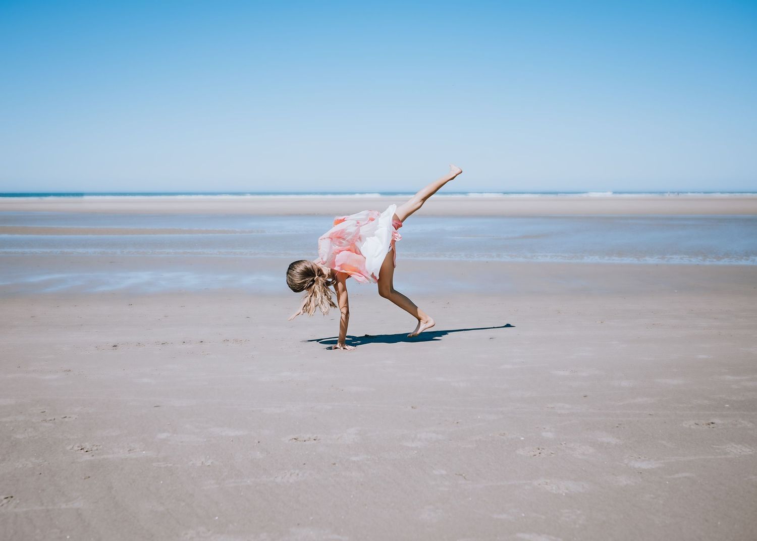 Figure doing acrobatic moves in pink dress on wet beach sand.