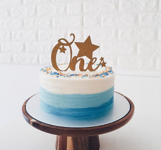 A blue ombre first birthday cake with gold 'One' topper and stars on a wooden cake stand.