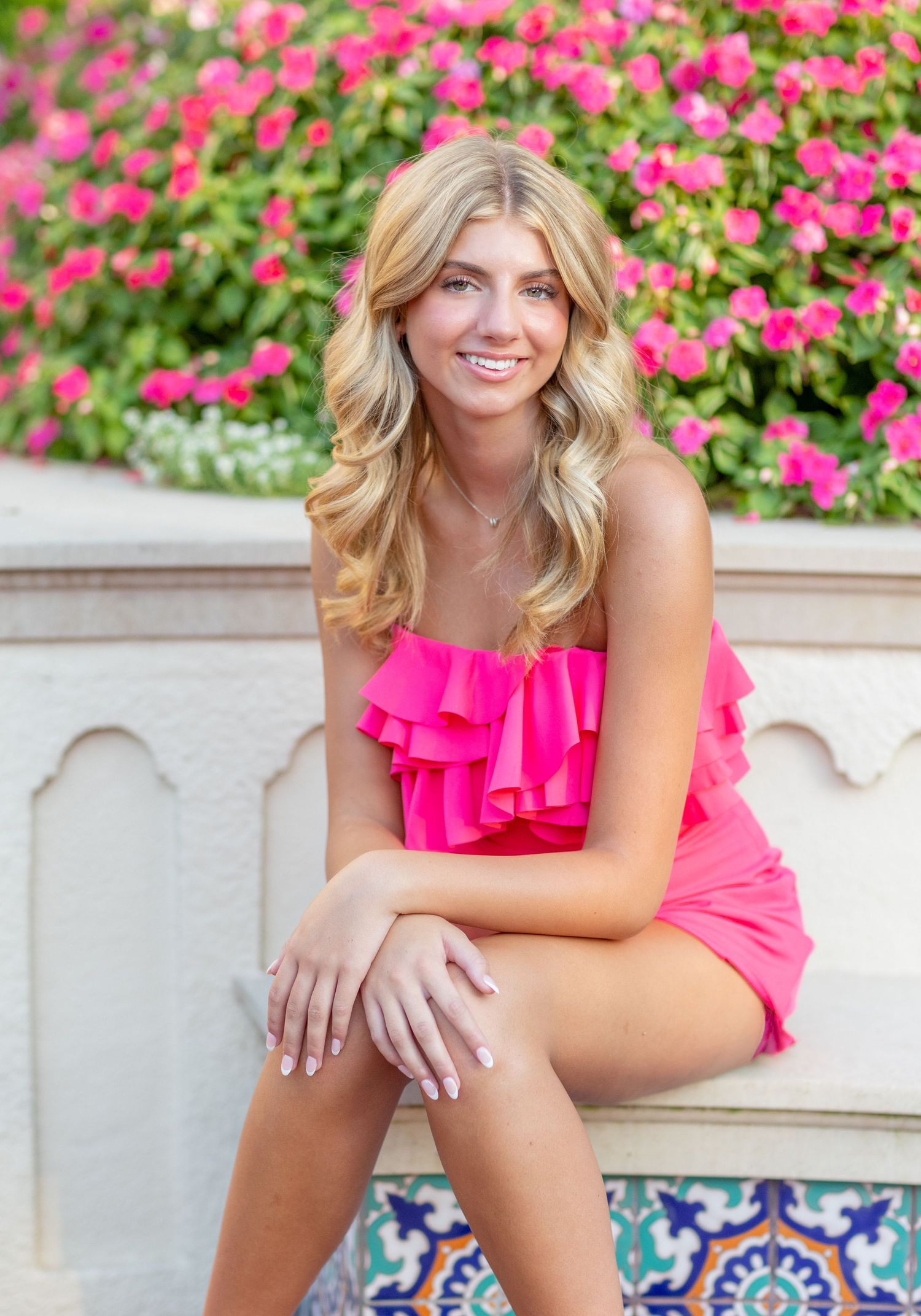 Young woman in bright pink top poses cheerfully among vibrant flower garden.