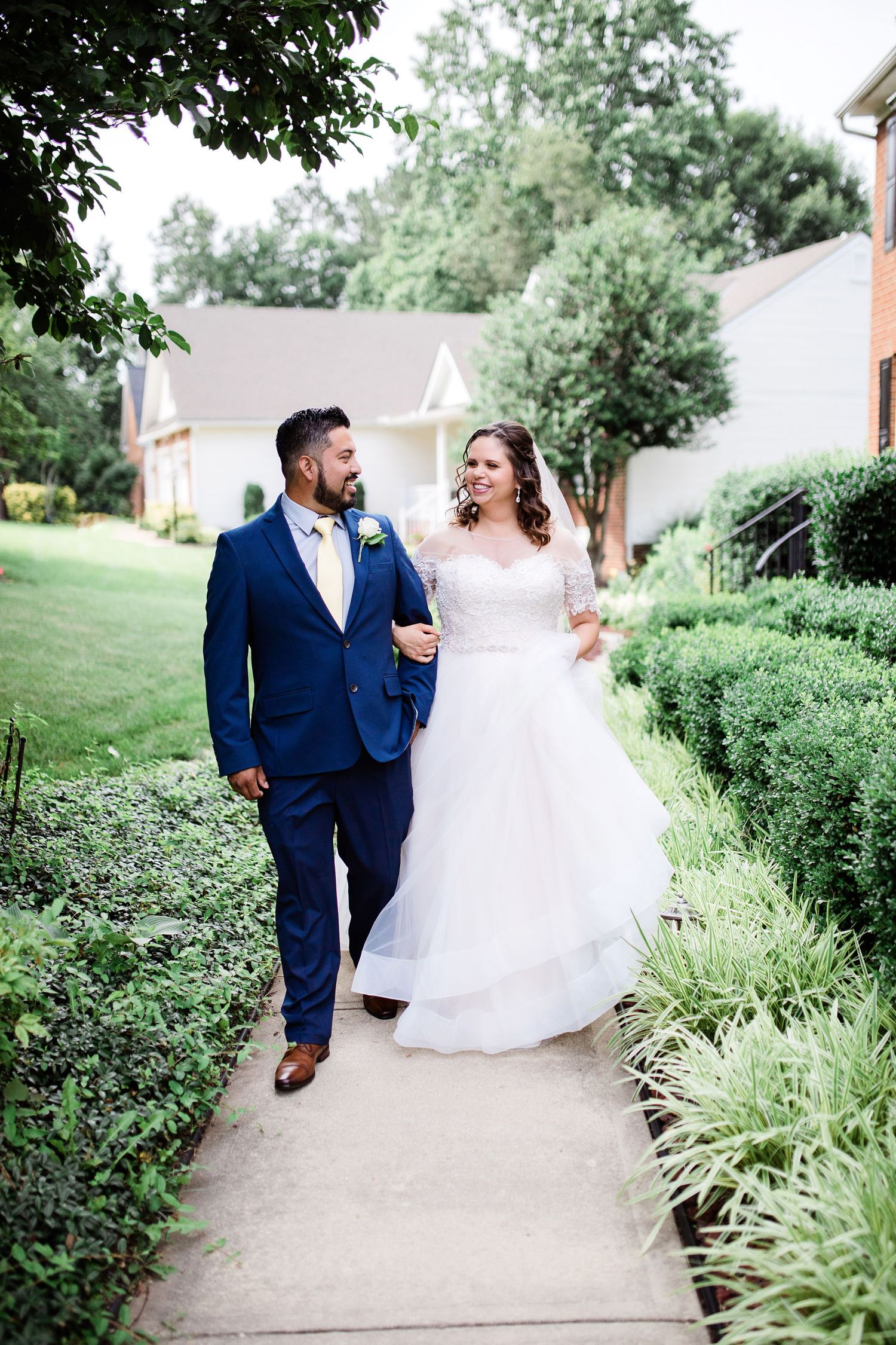 Newlywed couple walks hand in hand along garden path, groom in navy suit and bride in white gown.