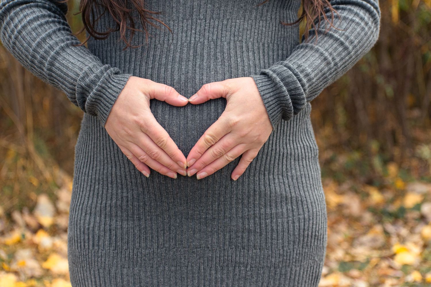 Hands forming a heart shape over a pregnant belly in a grey knit dress against an autumn background.