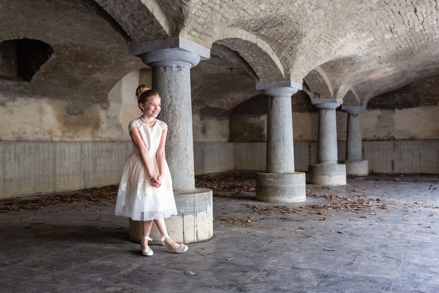 Child in white dress standing in historic stone basement with arched columns.