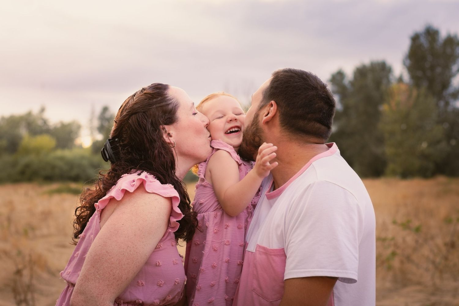 Mom and Dad kissing youngest sister on cheeks. Sister laughing. all matching outfits soft pink.