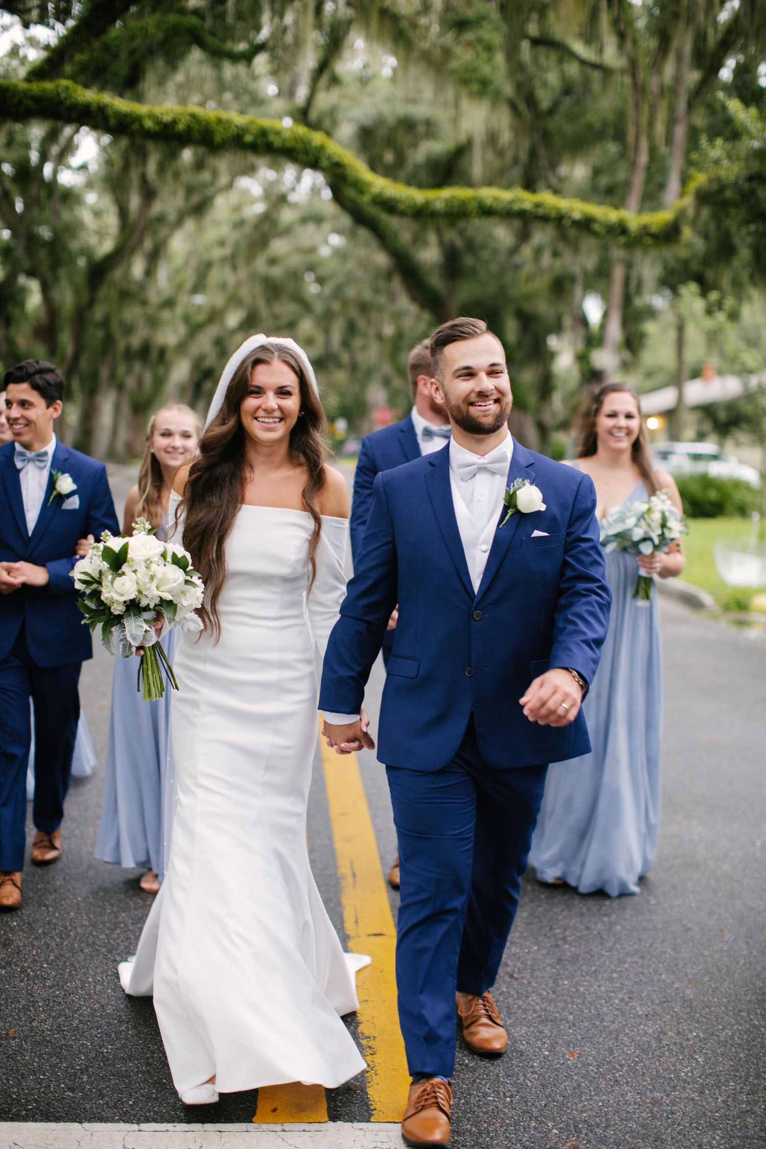 Wedding party walks down moss-covered tree lined street with bride in white and bridesmaids in blue.