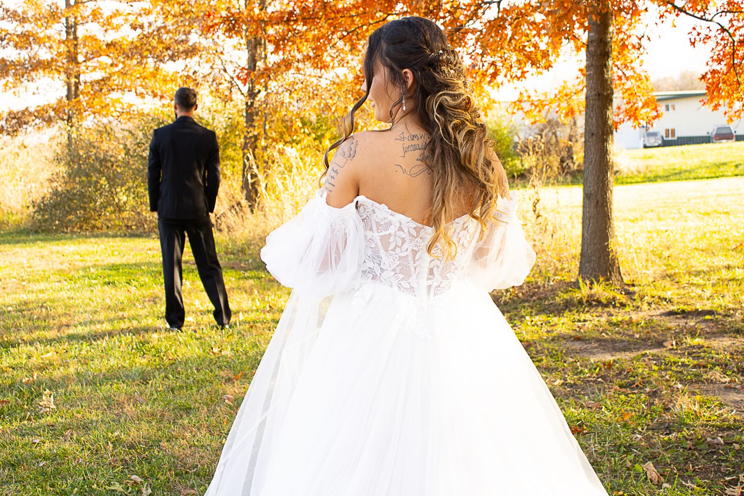 Back view of bride in lace dress with groom silhouetted against autumn trees.