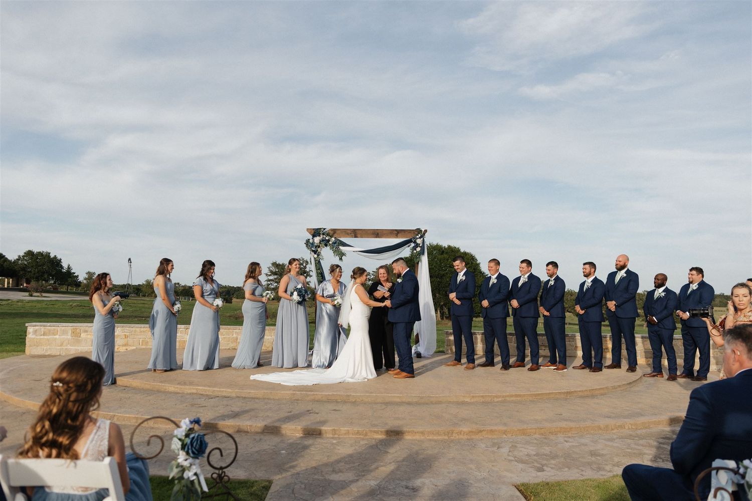 Wedding ceremony in progress under wooden arch with bridal party in navy and light blue.