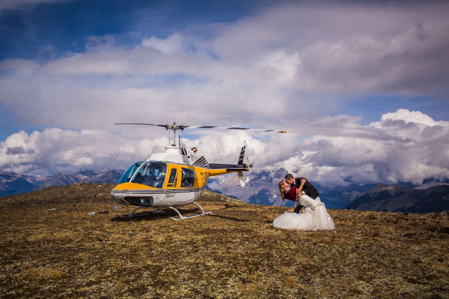 Wedding couple explores rugged mountain terrain with yellow helicopter in background.