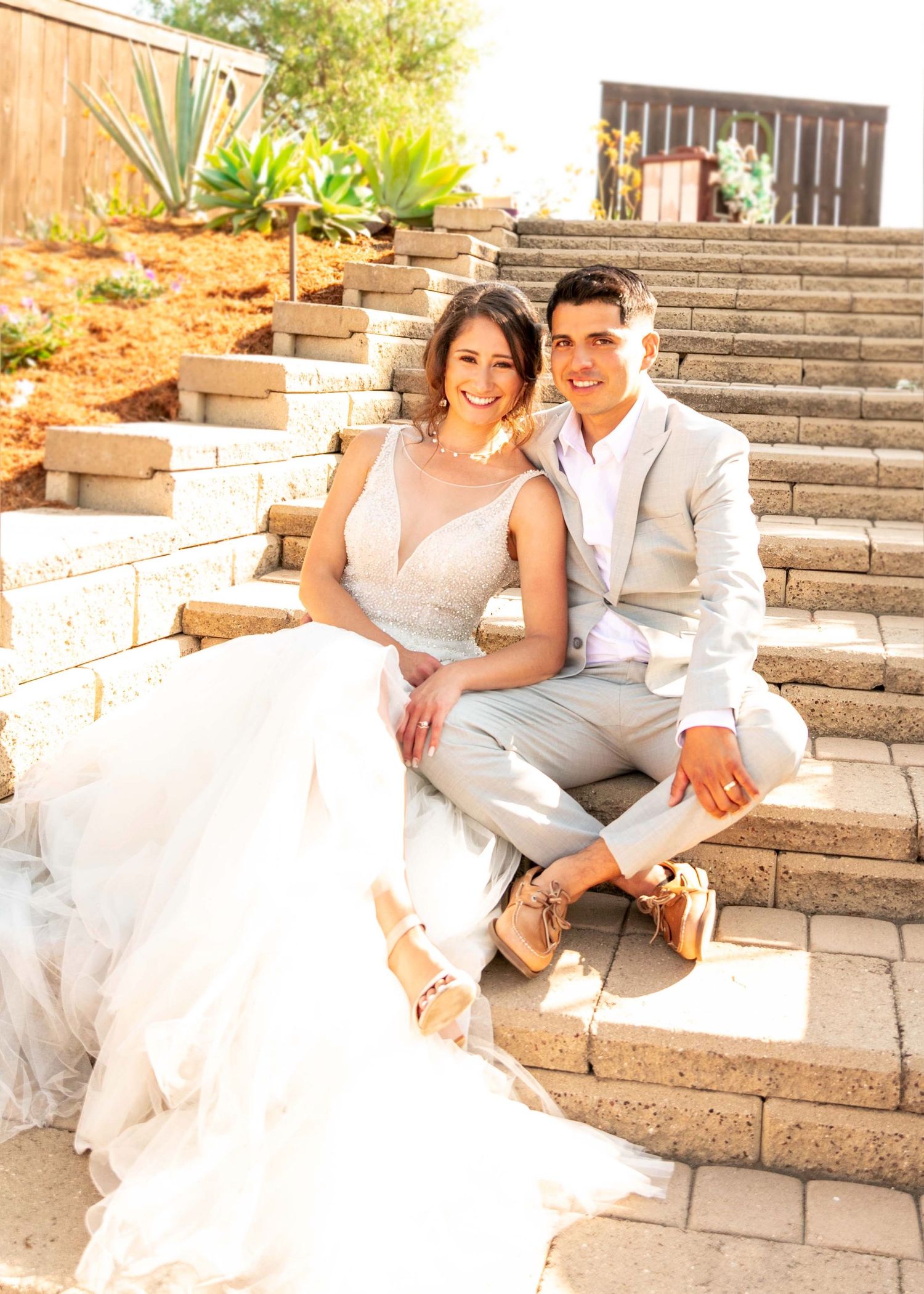 Wedding couple in formal attire sitting on outdoor stone steps surrounded by landscaping.