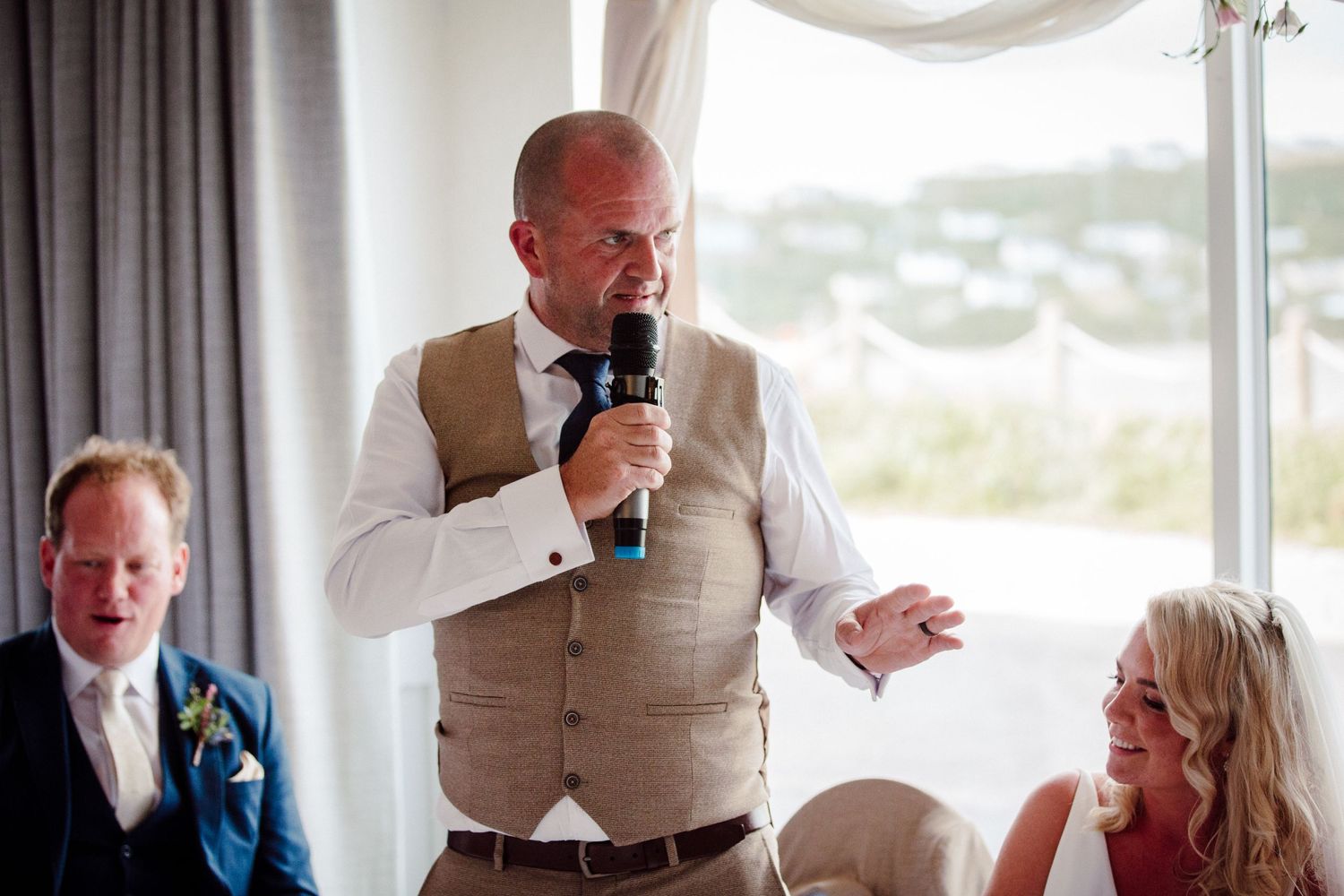 A speaker in a tan vest delivers a wedding speech while standing in a bright windowed room.