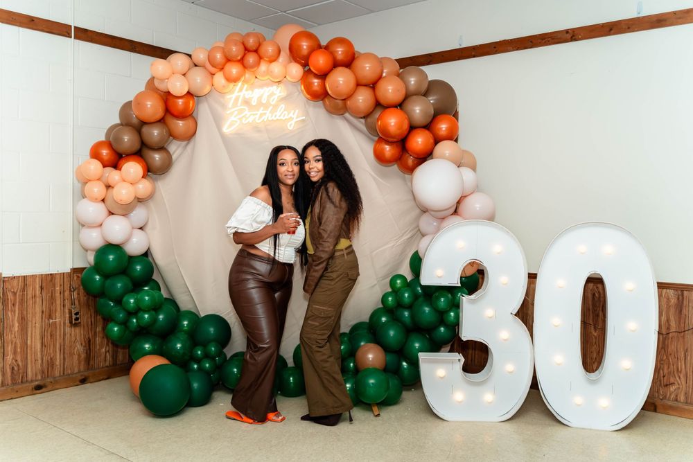 Two friends posing under decorative balloon arch with number 30 at a birthday celebration.